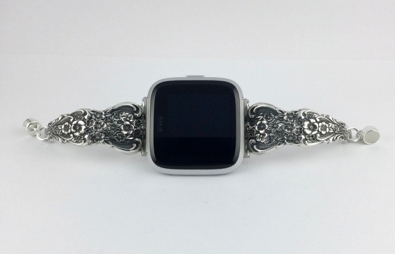 May include: Silver-toned Fitbit Versa smartwatch with a decorative floral band. The band features intricate floral and leaf patterns. The watch face is black, and the band connects with a clasp. The watch is displayed on a white background.