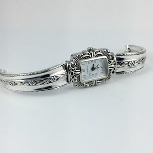 May include: A silver-toned watch with a square face and ornate detailing. The watch band is a silver-toned cuff bracelet with floral designs.