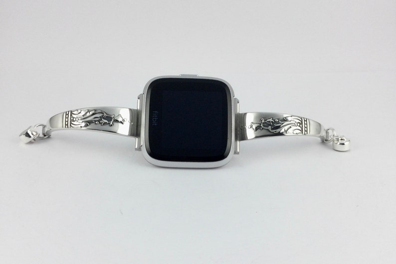 Op de afbeelding: Zilveren horlogeband met een sierlijk ontwerp, met een Fitbit smartwatch met een zwart scherm.