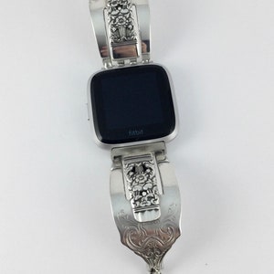 May include: A silver Fitbit smartwatch with a custom-made, ornate, silver spoon band. The band has a floral design and a dangling charm.