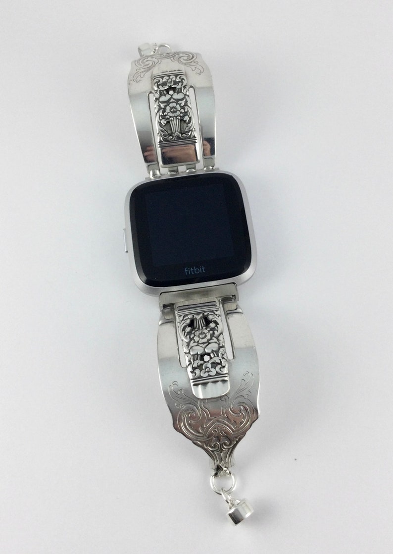 May include: A silver Fitbit smartwatch with a custom-made, ornate, silver spoon band. The band has a floral design and a dangling charm.