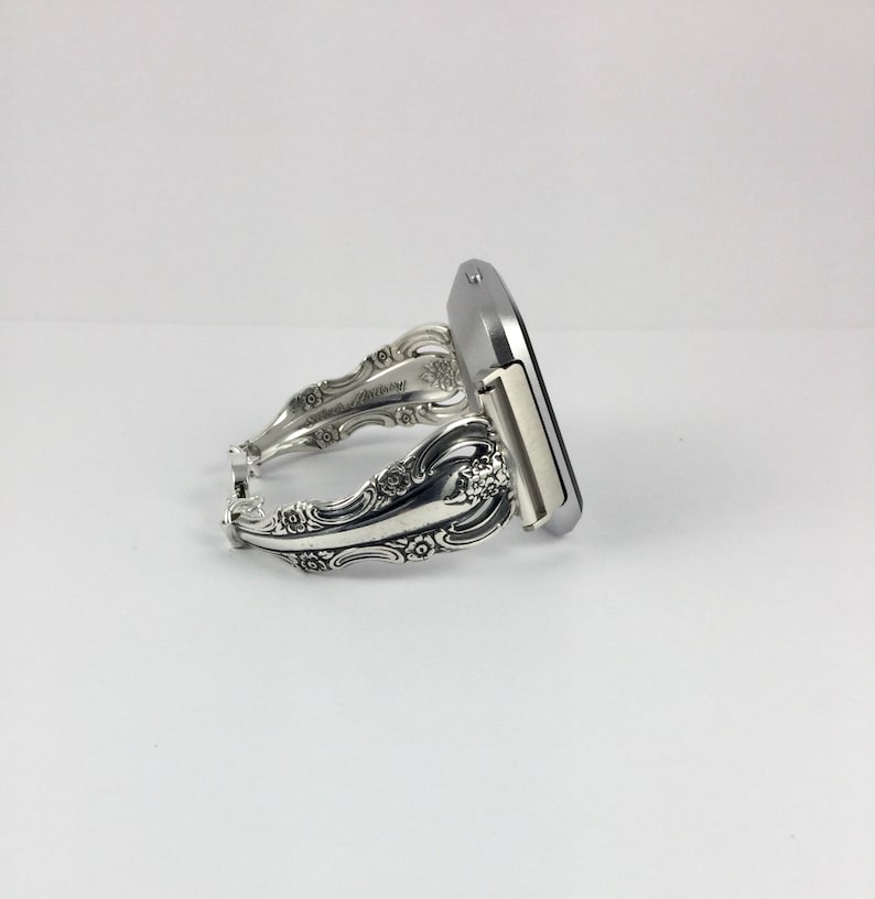 May include: A silver watch band made from a repurposed silverware piece. The band is decorated with floral designs and has a silver watch face.