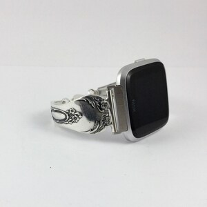 May include: Silver watch band made from a vintage silverware piece with floral details. The band is attached to a silver Fitbit smartwatch with a black screen.