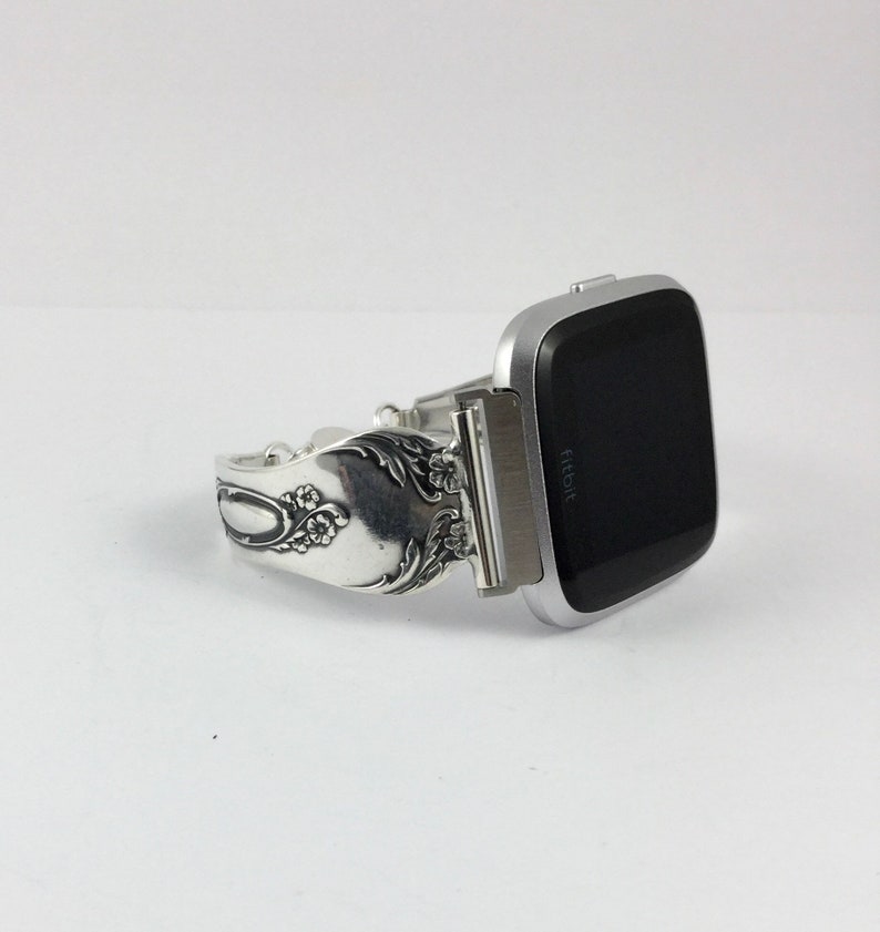 May include: Silver watch band made from a vintage silverware piece with floral details. The band is attached to a silver Fitbit smartwatch with a black screen.