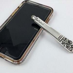 May include: A silver stylus with an ornate design on the handle, resting on a clear phone case.