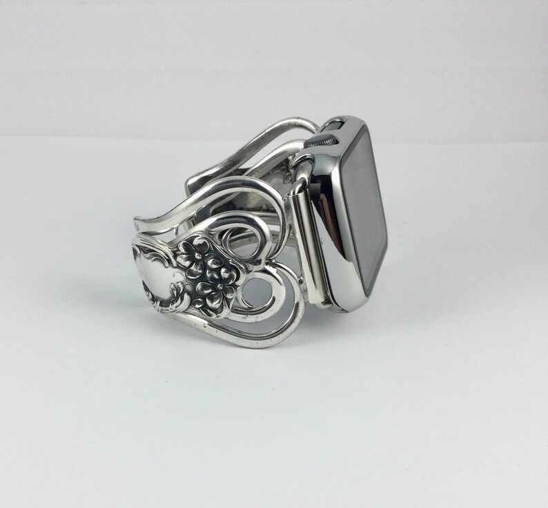 May include: Silver-toned watch band with a decorative floral and scroll design. The band is made from repurposed silverware, featuring spoon and fork elements. The watch face is a shiny, reflective black.
