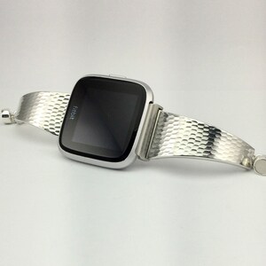 K&ouml;nnte beinhalten: Eine silberne Fitbit Smartwatch mit einem strukturierten silbernen Armband. Das Armband ist einteilig, umschlie&szlig;t das Handgelenk und verf&uuml;gt &uuml;ber einen magnetischen Verschluss.