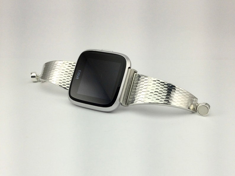 K&ouml;nnte beinhalten: Eine silberne Fitbit Smartwatch mit einem strukturierten silbernen Armband. Das Armband ist einteilig, umschlie&szlig;t das Handgelenk und verf&uuml;gt &uuml;ber einen magnetischen Verschluss.
