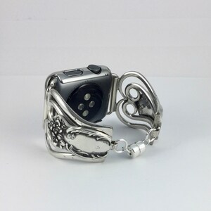 May include: Silver-toned watch band crafted from repurposed silverware. The band features floral and heart-shaped designs, with a magnetic clasp. The watch face is a dark gray color.