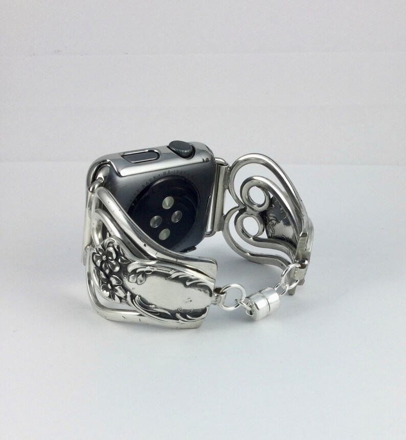 May include: Silver-toned watch band crafted from repurposed silverware. The band features floral and heart-shaped designs, with a magnetic clasp. The watch face is a dark gray color.