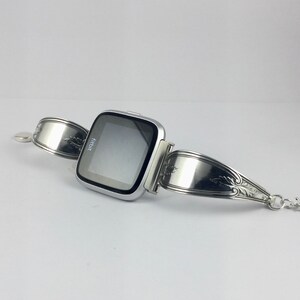 May include: A silver Fitbit smartwatch with a black screen displaying the word "fitbit" on a silver spoon bracelet.