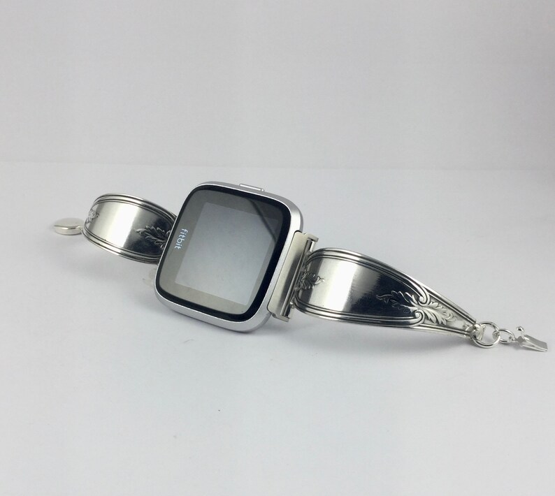 May include: A silver Fitbit smartwatch with a black screen displaying the word "fitbit" on a silver spoon bracelet.