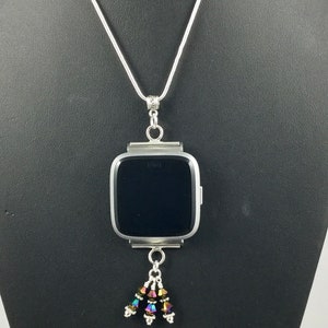 Puede incluir: Collar plateado con un colgante de reloj Fitbit. El reloj tiene una pantalla negra y est&aacute; adornado con un grupo de cuentas iridiscentes. El collar se exhibe en un maniqu&iacute; negro.
