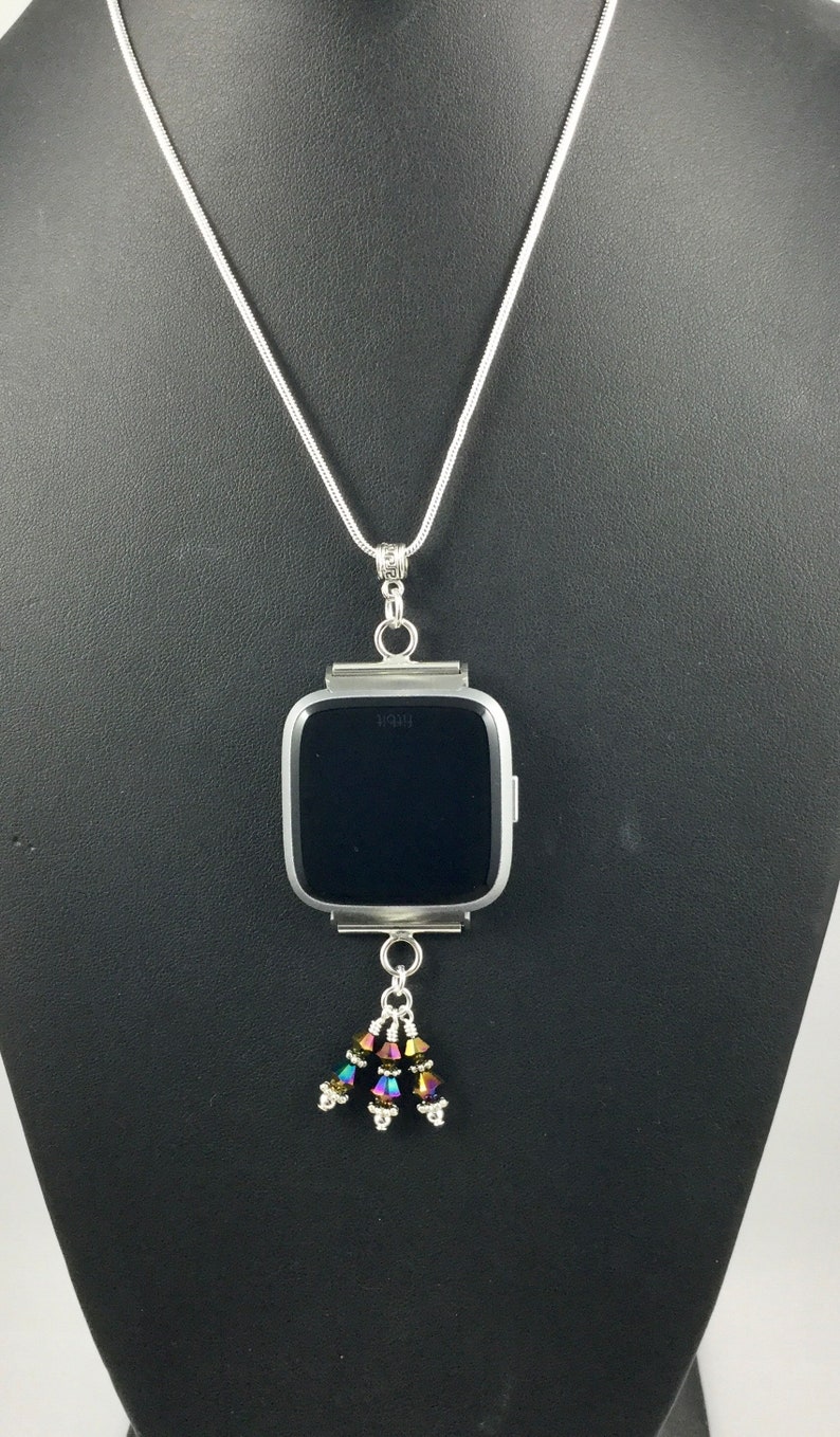 Puede incluir: Collar plateado con un colgante de reloj Fitbit. El reloj tiene una pantalla negra y est&aacute; adornado con un grupo de cuentas iridiscentes. El collar se exhibe en un maniqu&iacute; negro.