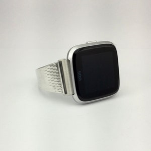 K&ouml;nnte beinhalten: Eine silberne Fitbit Smartwatch mit einem strukturierten Metallband. Das Zifferblatt der Uhr ist schwarz und zeigt das Wort "fitbit" an.
