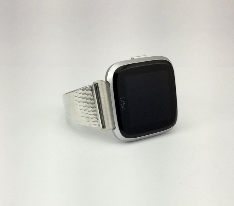 K&ouml;nnte beinhalten: Eine silberne Fitbit Smartwatch mit einem strukturierten Metallband. Das Zifferblatt der Uhr ist schwarz und zeigt das Wort "fitbit" an.