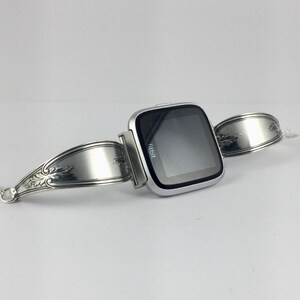May include: A silver Fitbit smartwatch attached to a silver bracelet made from a repurposed spoon.