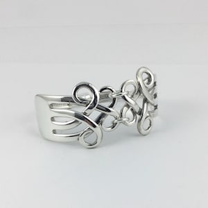 May include: A silver-toned bracelet crafted from a fork, featuring a decorative Celtic knot design. The bracelet has a fork head on one end and intricate, swirling patterns on the other, creating a unique and artistic piece of jewelry.