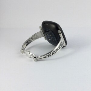 Puede incluir: Un brazalete de plata con un dise&ntilde;o intrincado, que sostiene un reloj inteligente Fitbit negro. El reloj inteligente tiene un sensor de frecuencia card&iacute;aca en la parte posterior.