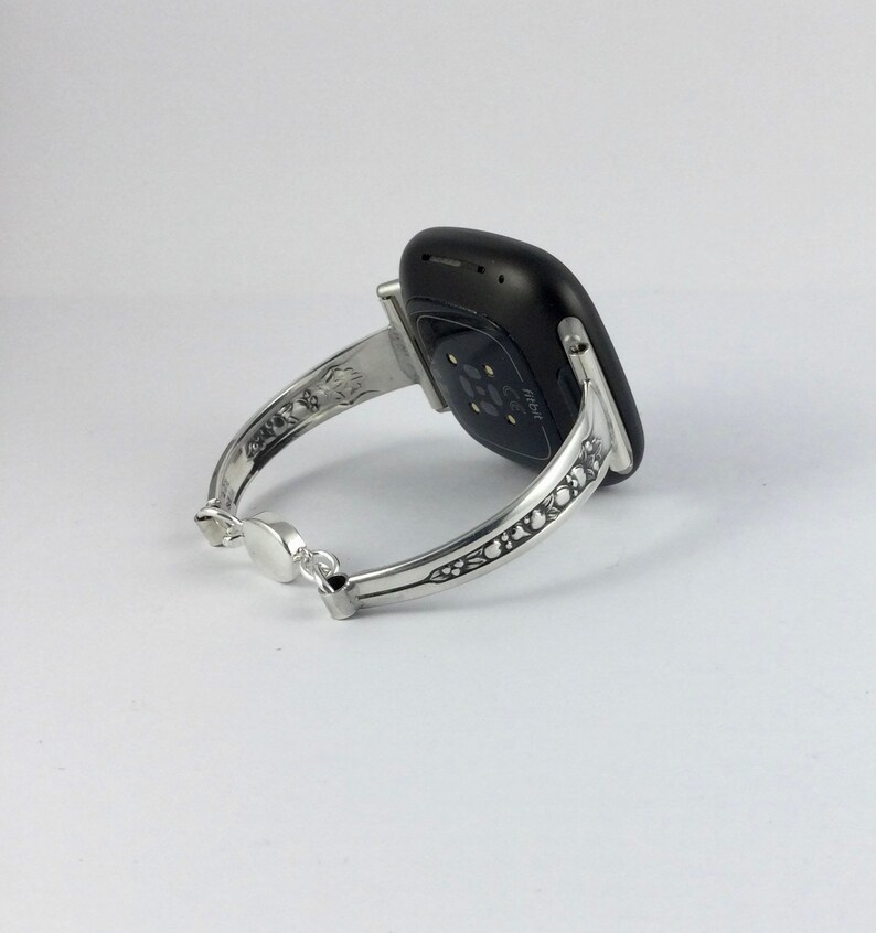 Puede incluir: Un brazalete de plata con un dise&ntilde;o intrincado, que sostiene un reloj inteligente Fitbit negro. El reloj inteligente tiene un sensor de frecuencia card&iacute;aca en la parte posterior.