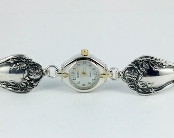 Könnte beinhalten: Eine silberfarbene Quarzuhr mit weißem Zifferblatt, schwarzen Ziffern und einer goldfarbenen Krone. Das Armband besteht aus zwei verzierten, silberfarbenen Löffelgriffen mit geprägten Rosenmotiven, die durch Metallglieder verbunden sind.