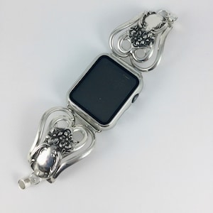 May include: Silver-toned watch band with a rectangular watch face. The band features a floral design with heart-shaped links. The watch face is black. The band is made of metal and has a vintage aesthetic.