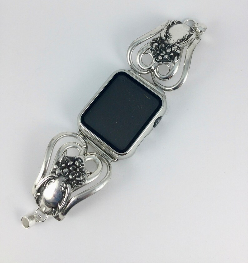 May include: Silver-toned watch band with a rectangular watch face. The band features a floral design with heart-shaped links. The watch face is black. The band is made of metal and has a vintage aesthetic.