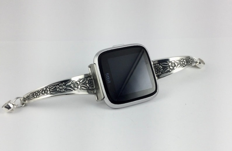 Puede incluir: Un reloj inteligente Fitbit plateado con una banda con estampado floral. La banda est&aacute; hecha de metal y tiene un cierre de broche.