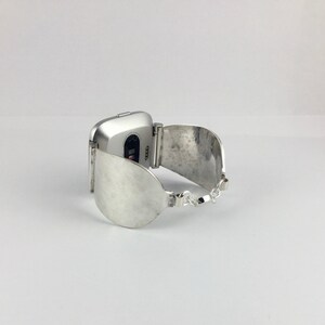 May include: Silver watch band with a curved, silver cuff design. The watch face is square and silver.