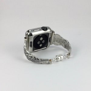 May include: Silver-toned Apple Watch with a decorative band. The band features an ornate floral design and a magnetic clasp. The watch case is silver and has a black button. The watch is shown on a white background.