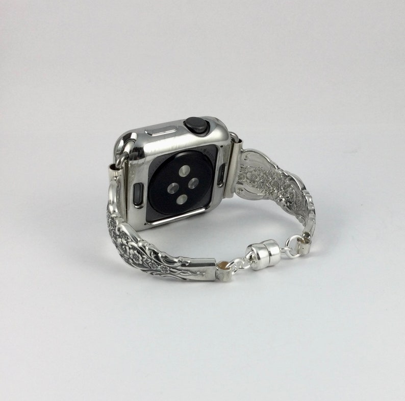 May include: Silver-toned Apple Watch with a decorative band. The band features an ornate floral design and a magnetic clasp. The watch case is silver and has a black button. The watch is shown on a white background.