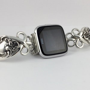 May include: A silver Fitbit smartwatch with a black screen is attached to a silver bracelet made of intricately designed spoons.