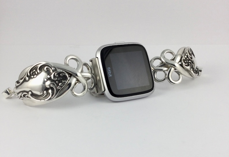 May include: A silver Fitbit smartwatch with a black screen is attached to a silver bracelet made of intricately designed spoons.