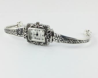May include: Silver-toned watch with a rectangular face and ornate floral detailing. The watch features a white dial with black numerals and hands. The band is a cuff style with matching floral designs.