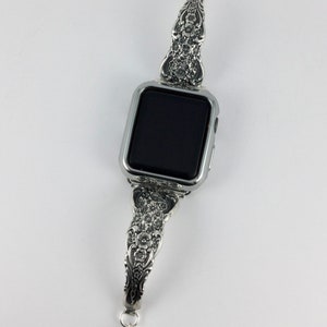 May include: Silver-toned watch band with ornate floral detailing. The band is designed to fit a smartwatch, featuring a rectangular watch face. The band's design includes a small, dangling charm at the clasp. The watch face is black.