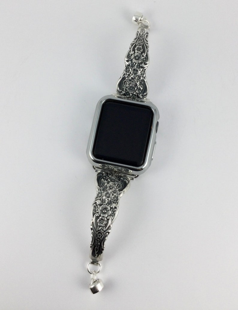 May include: Silver-toned watch band with ornate floral detailing. The band is designed to fit a smartwatch, featuring a rectangular watch face. The band's design includes a small, dangling charm at the clasp. The watch face is black.