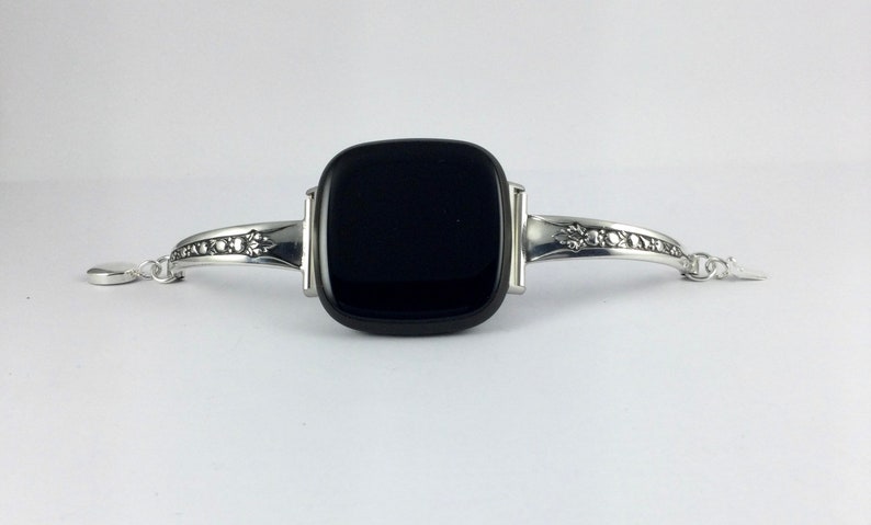 Puede incluir: Una pulsera de plata con una piedra negra cuadrada en el centro. La pulsera tiene una cadena delicada y dise&ntilde;os florales en los lados.