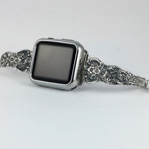 May include: Silver-toned Apple Watch band with ornate floral detailing. The band features a decorative design on both sides of the watch face and a magnetic clasp. The watch face is a dark gray color.