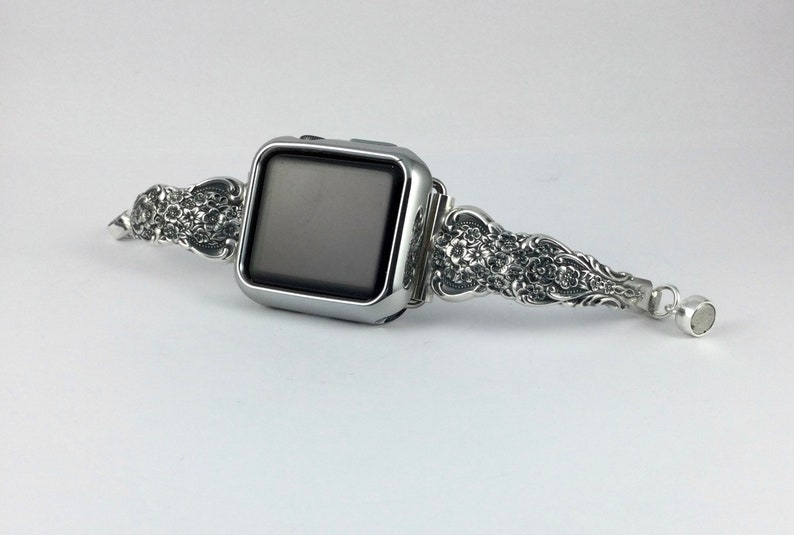 May include: Silver-toned Apple Watch band with ornate floral detailing. The band features a decorative design on both sides of the watch face and a magnetic clasp. The watch face is a dark gray color.