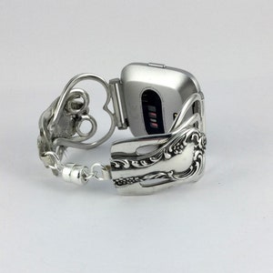 Puede incluir: Pulsera de reloj de plata hecha de una cuchara reutilizada con un dise&ntilde;o en forma de coraz&oacute;n. La pulsera est&aacute; unida a un reloj inteligente plateado.