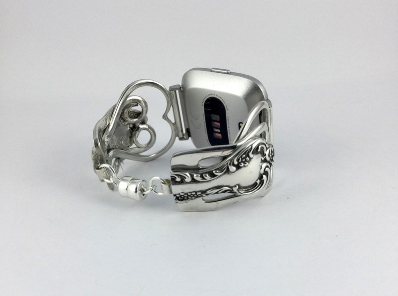 Puede incluir: Pulsera de reloj de plata hecha de una cuchara reutilizada con un dise&ntilde;o en forma de coraz&oacute;n. La pulsera est&aacute; unida a un reloj inteligente plateado.