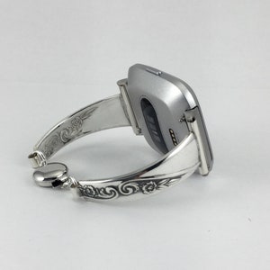 May include: A silver watch band with an ornate design. The band is made of metal and has a floral pattern. The watch band is designed to fit a square-shaped smartwatch.