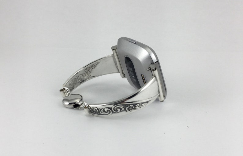 May include: A silver watch band with an ornate design. The band is made of metal and has a floral pattern. The watch band is designed to fit a square-shaped smartwatch.