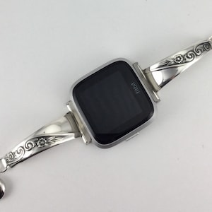 May include: Silver watch band with an ornate design featuring floral details. The band is attached to a square smartwatch with a black screen that displays the word "Fitbit".