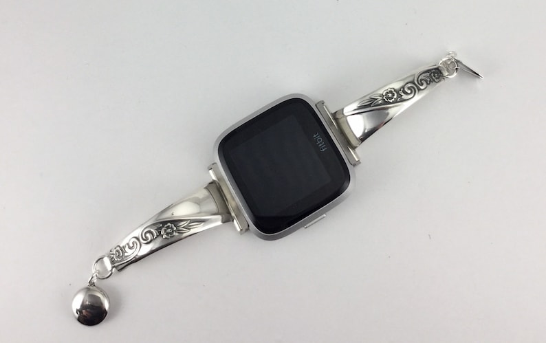 May include: Silver watch band with an ornate design featuring floral details. The band is attached to a square smartwatch with a black screen that displays the word "Fitbit".
