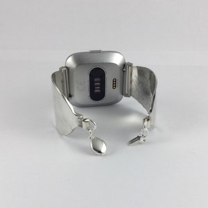 May include: Silver smartwatch with a silver metal band. The watch face is square and has a black screen with a white CE mark and four small black dots.