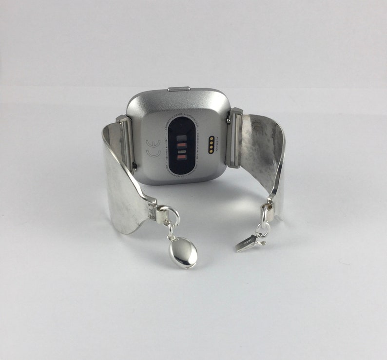 May include: Silver smartwatch with a silver metal band. The watch face is square and has a black screen with a white CE mark and four small black dots.