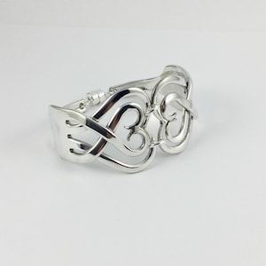 May include: A silver bracelet with a heart design. The bracelet is made of intertwined heart shapes and has a fork-like clasp. The bracelet is on a white background.