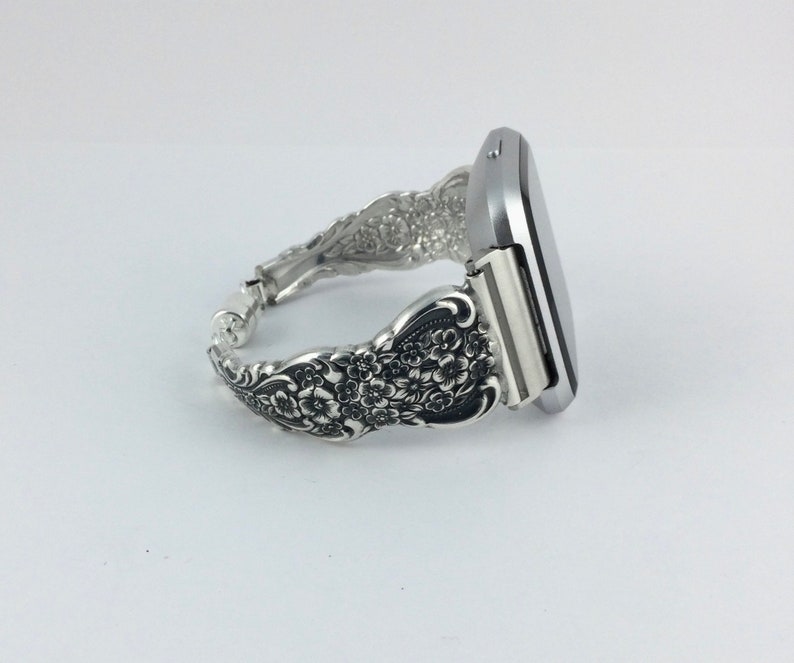 May include: A silver-toned watch band crafted from ornate silverware. The band features a floral design and connects to a silver watch face. The watch band is a unique accessory, combining functionality with vintage style.