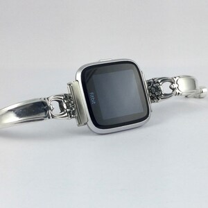 May include: A silver Fitbit smartwatch with a black screen is attached to a silver bracelet made from a repurposed silverware handle.